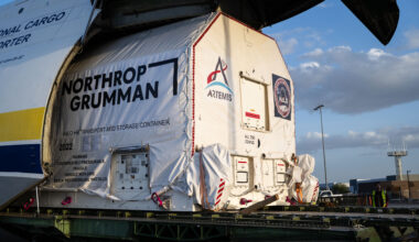 A large cargo aircraft with its nose lifted open reveals a wrapped container being offloaded. The container is marked with the logo for NASA's Artemis campaign and labeled as the HALO transport and storage container. The scene is set at an airport during late afternoon.