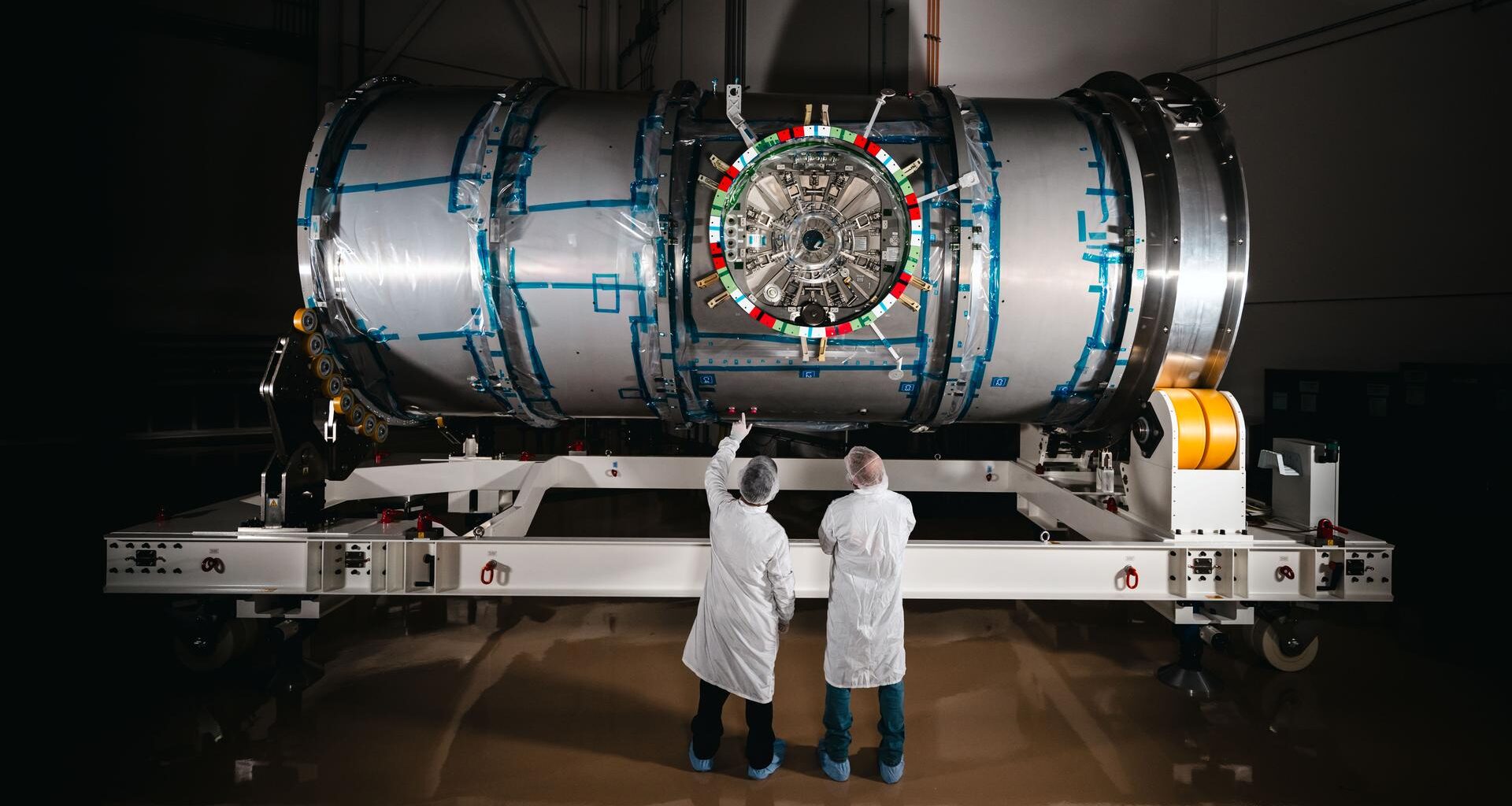 Two technicians in cleanroom attire stand in front of HALO (Habitation and Logistics Outpost) at Northrop Grumman’s facility in Gilbert, Arizona. The cylindrical metallic module, recently unboxed, rests on a white stand. One technician points toward the front of the module.