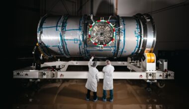Two technicians in cleanroom attire stand in front of HALO (Habitation and Logistics Outpost) at Northrop Grumman’s facility in Gilbert, Arizona. The cylindrical metallic module, recently unboxed, rests on a white stand. One technician points toward the front of the module.