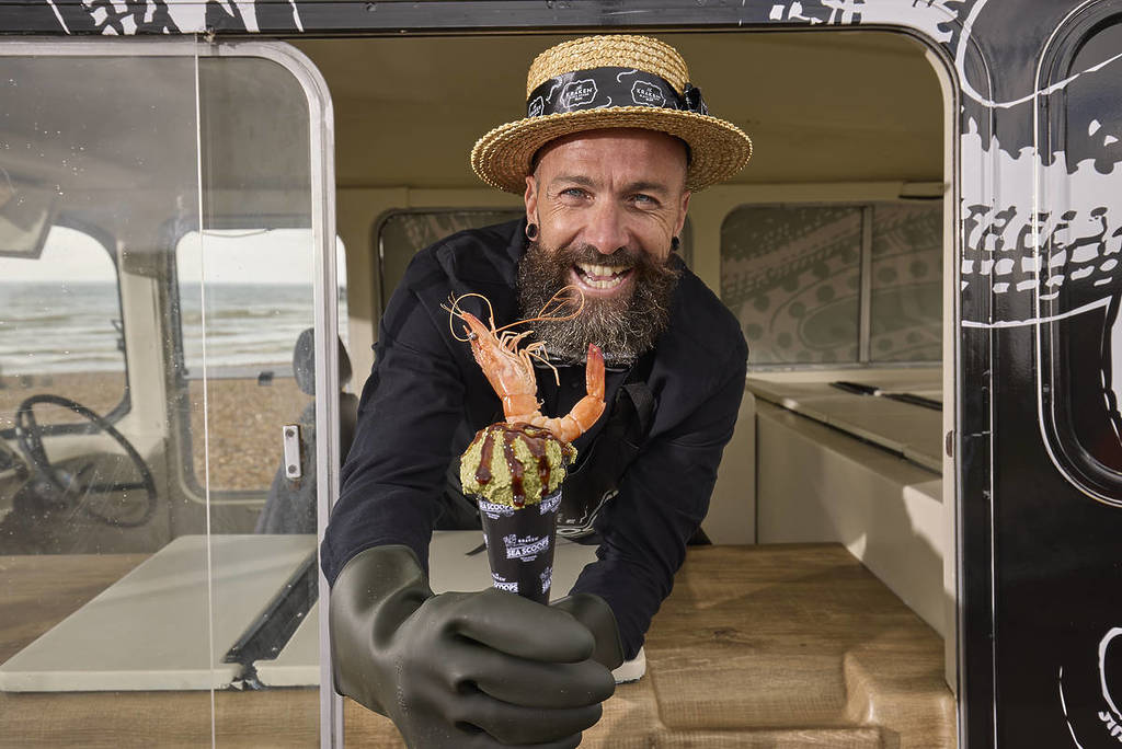 man holding taramasalats coop with king prawn on top from ice cream-style van and wearing straw boater
