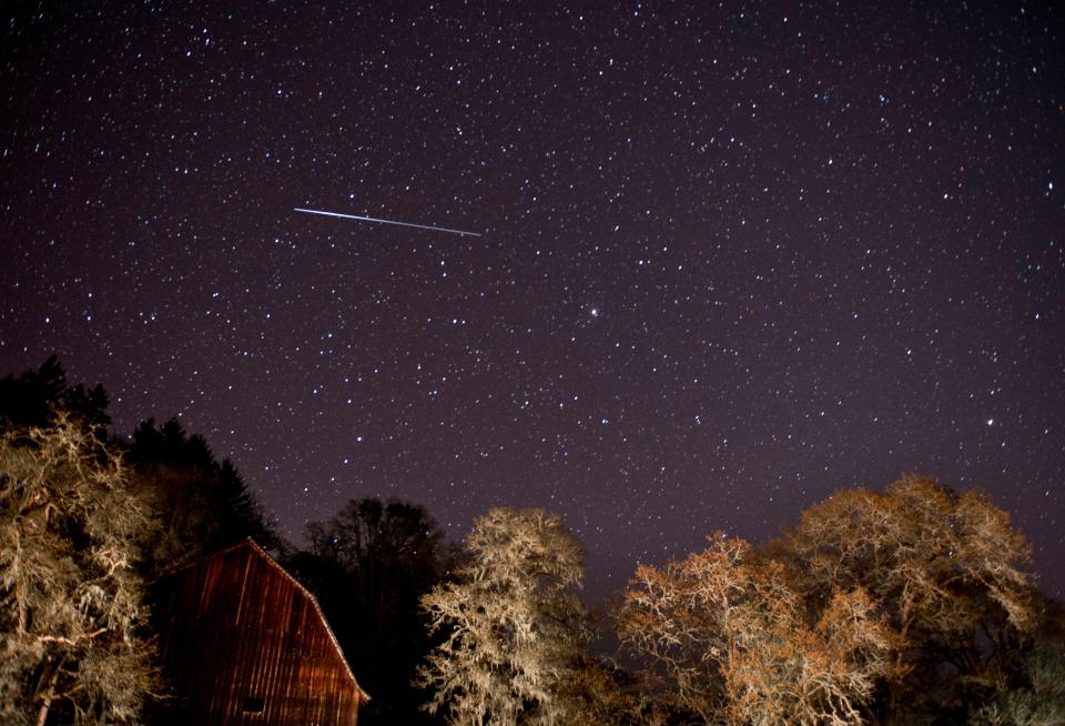 Meteor shower streaks across a night sky above a barn and trees.