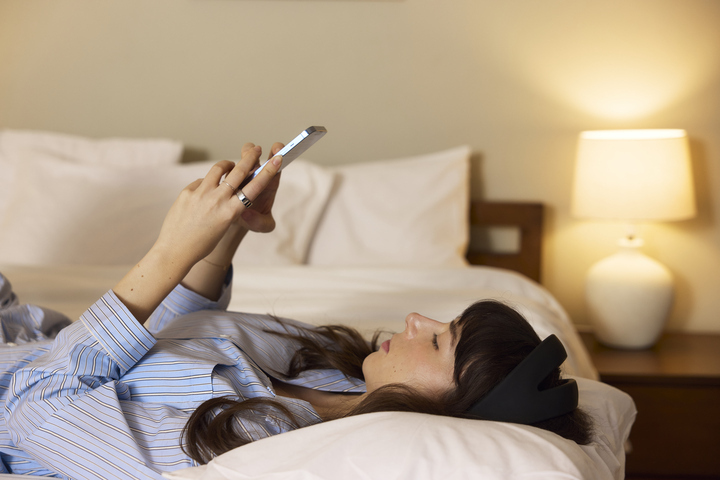 Woman lying in bed using a smartphone while wearing a headband.
