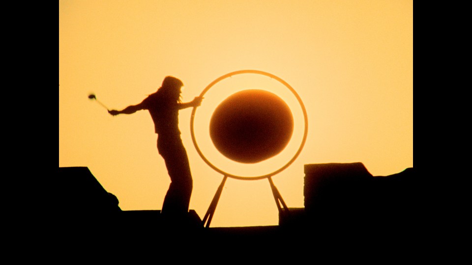 Silhouette of a person playing a large gong at sunset.