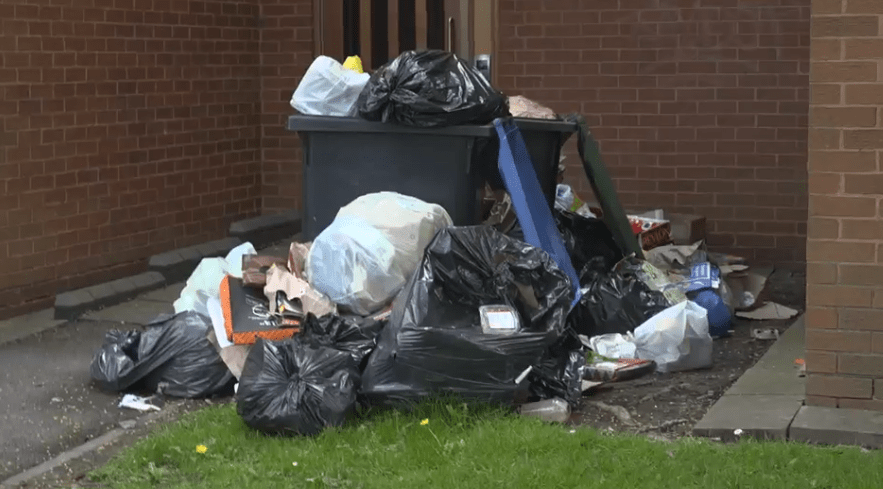 Overflowing trash bags and a full dumpster outside a brick building.