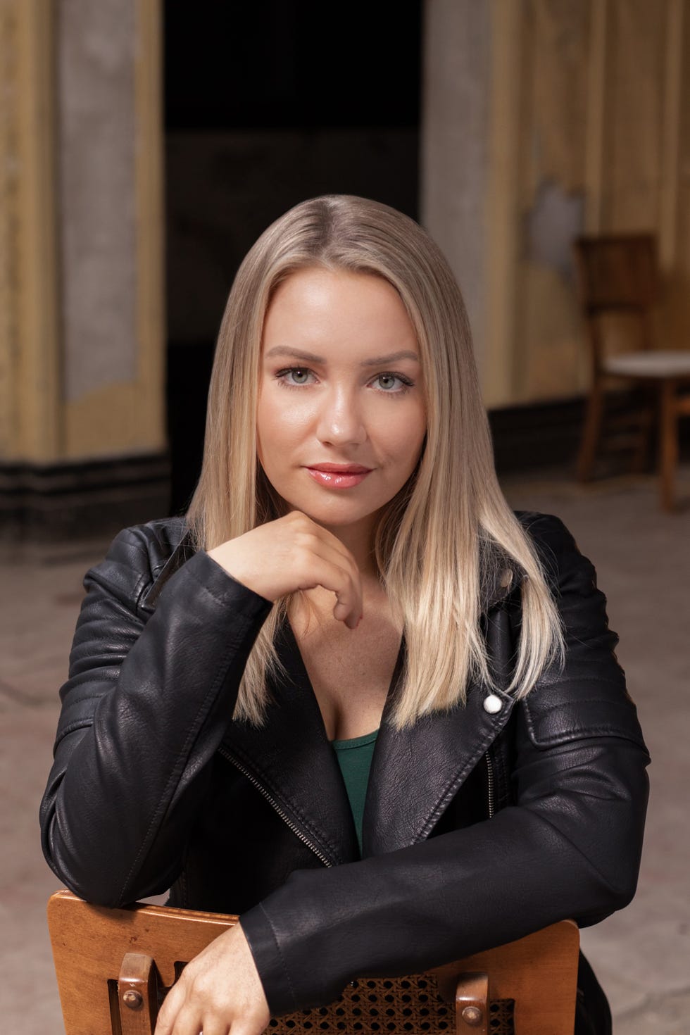 a seated individual with long blonde hair wearing a black leather jacket resting their chin on their hand