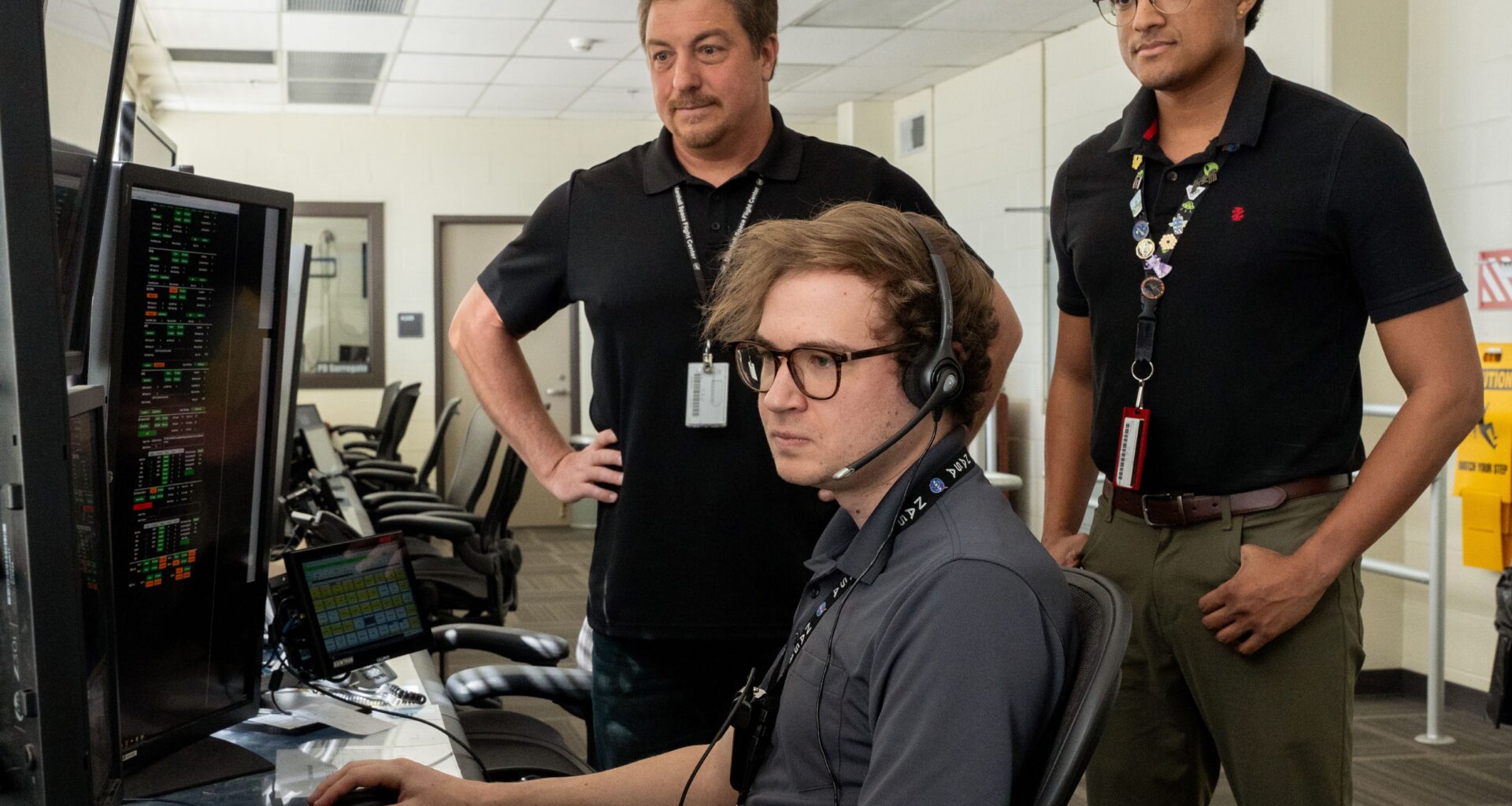 Three men standing inside a computer room reviewing information on a computer screen.