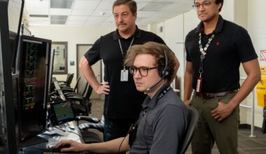 Three men standing inside a computer room reviewing information on a computer screen.