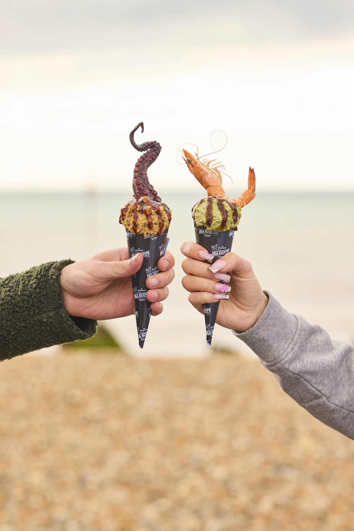 two seafood soft serve scoop cones in hands on beach - left one with octopus topping, right one with king prawn topping