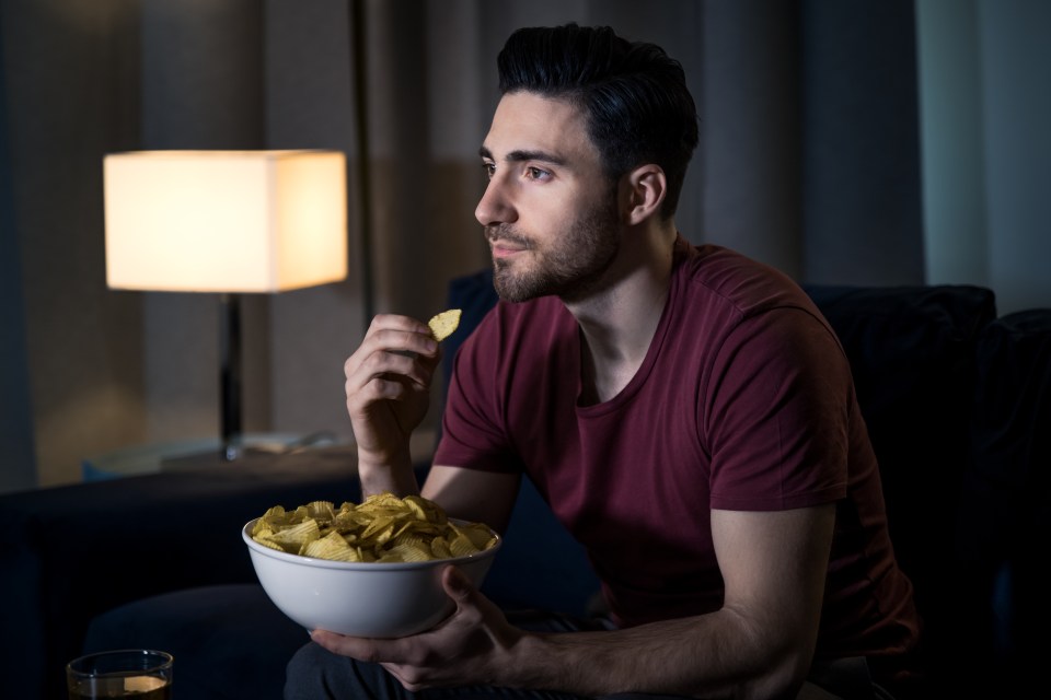 A young man relaxing on a sofa, eating potato chips while watching a movie.