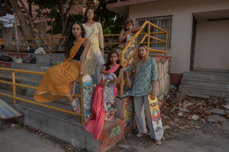 Five women wearing colorful saris pose with skateboards on an urban stairway, surrounded by yellow railings and concrete, with trees and a building in the background.