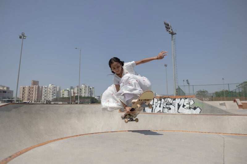 A girl in a white dress performs a skateboard trick in a skatepark, with buildings, graffiti, and a clear blue sky in the background.