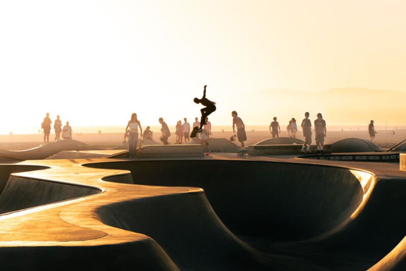 A skateboarder performs a trick in mid-air at a skatepark during sunset, with silhouettes of people walking and watching in the background. The scene is bathed in golden light.