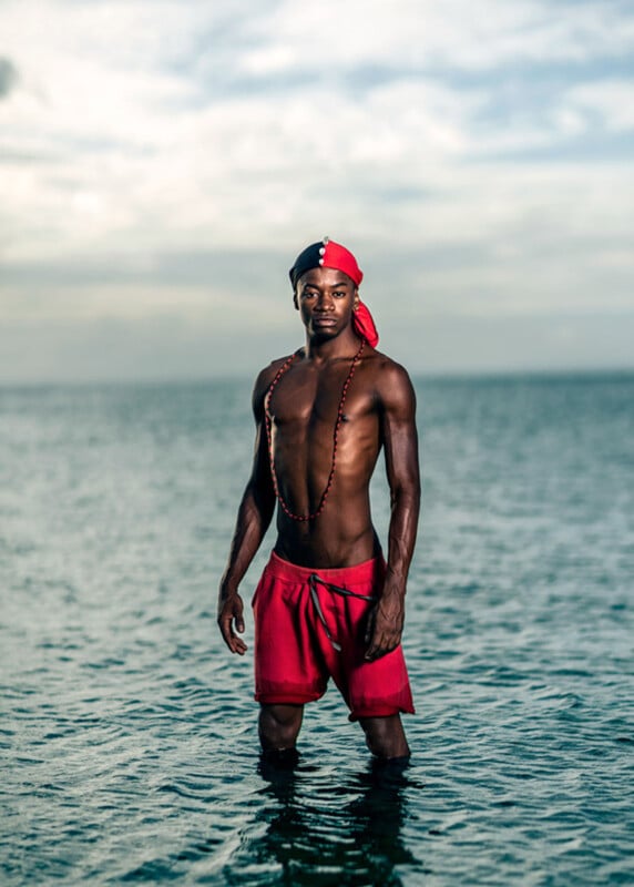 A man wearing red shorts, a red and black headscarf, and a necklace stands waist-deep in calm ocean water under a cloudy sky.