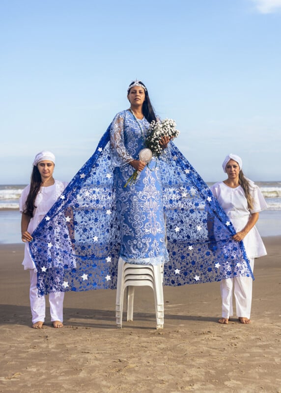 A woman in a long, blue, patterned dress and crown stands on stacked chairs at the beach, holding flowers, flanked by two women in white outfits holding her cape, with the ocean and sky in the background.