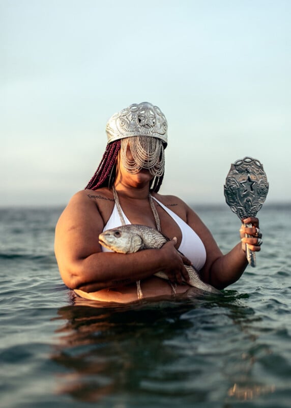 A person in a white bikini stands waist-deep in water, holding a fish and an ornate handheld mirror. They wear a decorative silver headdress with chains covering their face. The sky is clear in the background.