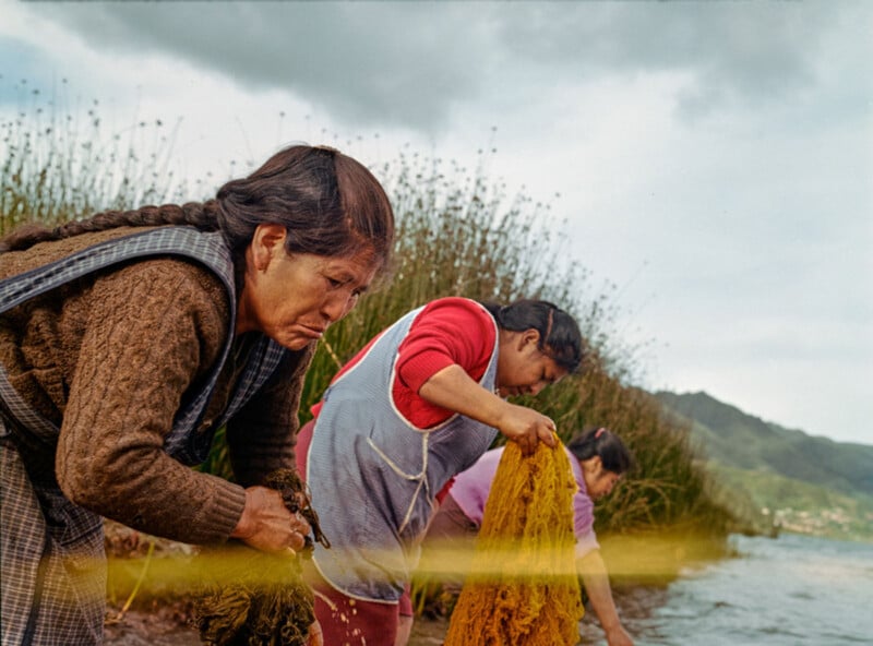 Three women stand at the edge of a lake, washing fabrics and plants by hand. Tall reeds grow nearby, and mountains are visible under a cloudy sky in the background. The women appear focused on their work.