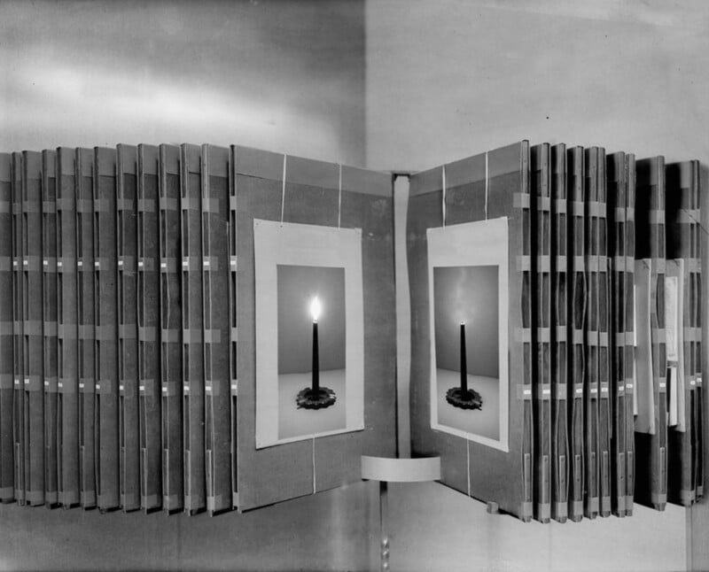 A black and white photo shows rows of file folders or books arranged in a semicircle, each with a photo of a lit candle on the front. The setup appears organized and symmetrical against a plain wall.