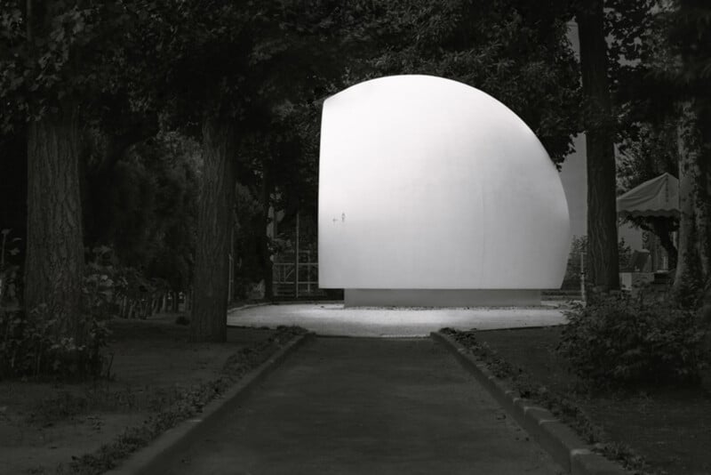 A large, smooth, white, dome-like structure stands in a park surrounded by tall trees and greenery, with a pathway leading directly toward it.