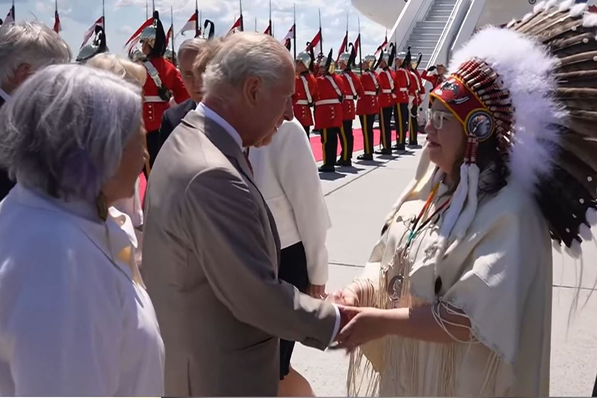 King Charles III shaking hands with an Indigenous woman in ceremonial headdress.