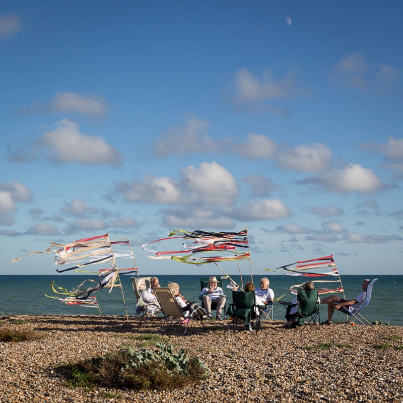 A group of people sit in camping chairs on a pebble beach facing the sea, surrounded by colorful windsocks fluttering in the breeze under a blue sky with scattered clouds.