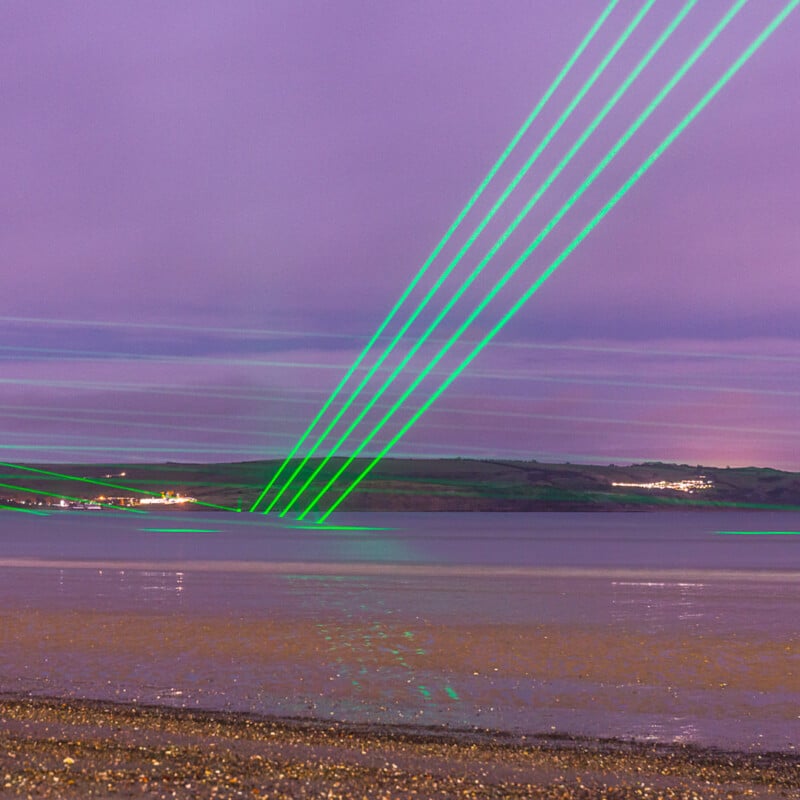 Green laser beams shoot diagonally across a purple sky above a calm body of water, with distant hills and lights visible on the horizon. The foreground shows a sandy shoreline at dusk or night.