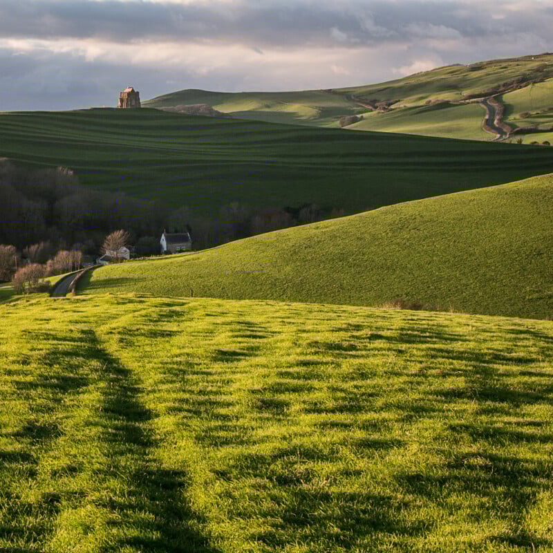 Rolling green hills under a cloudy sky, with a winding road, scattered houses, and a solitary stone building atop a distant hill. Sunlight illuminates the vibrant grass in the foreground.