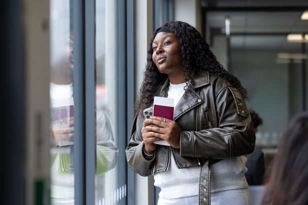 Waist up front view of a young female tourist waiting in the airport departure lounge ready for her flight. She is wearing casual clothing and she is looking out the window, she has her boarding pass, passport and smartphone in her hand.