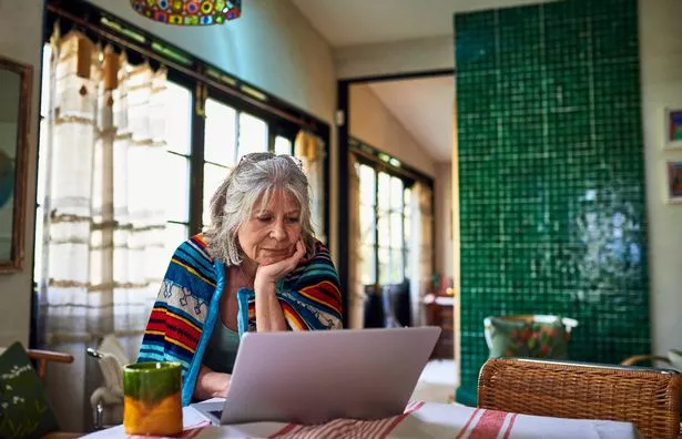 Senior woman wrapped in blanket standing at dining table looking at emails on laptop, morning, concentration, working from home