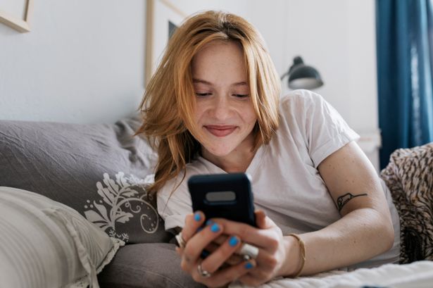 A woman smiling while lying on her bed at home and messaging on an online dating app using her smartphone.