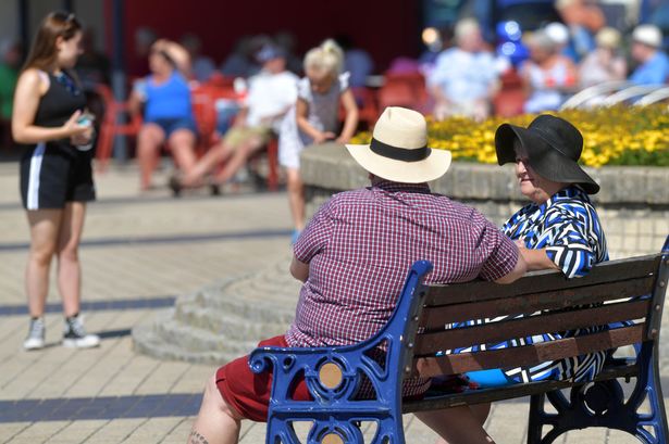 Two people sit on a bench in sunny weather wearing summer clothing 