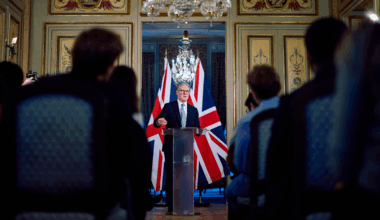 Keir Starmer, a man with grey hair and black glasses, wearing a blue tie and black suit, speaks at a podium in front of two UK flags. The room is ornate; there is a chandelier. Photo: Benjamin Cremel/Pool Photo via AP