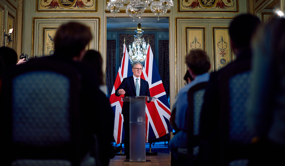 Keir Starmer, a man with grey hair and black glasses, wearing a blue tie and black suit, speaks at a podium in front of two UK flags. The room is ornate; there is a chandelier. Photo: Benjamin Cremel/Pool Photo via AP