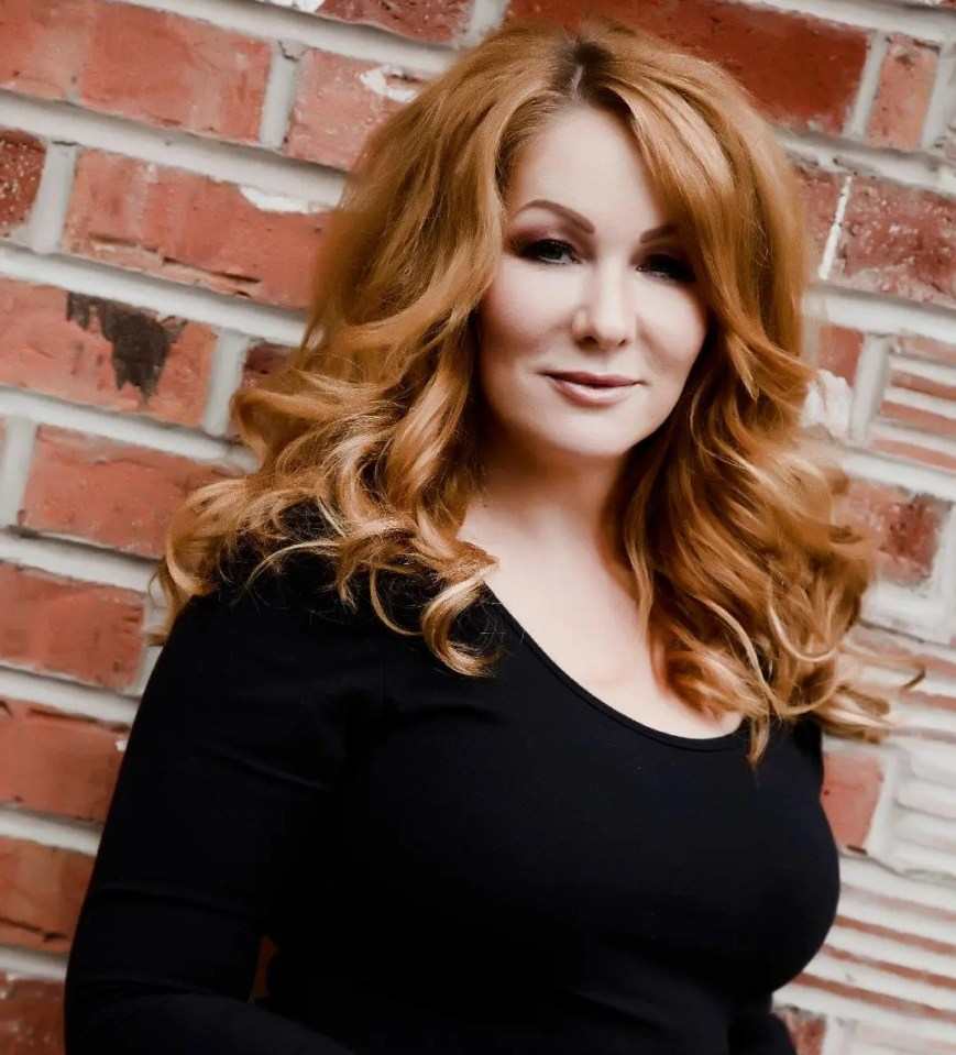 Woman with long red hair wearing a black dress against a brick wall.