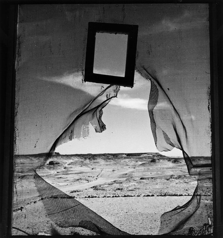 A torn mesh screen frames a barren desert landscape. A rectangular mirror or frame hangs in the sky, reflecting nothing. The scene is in black and white, with dramatic, surreal composition and strong contrasts.