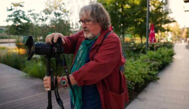 A person in a worn coat and backpack crouches on a city street, aiming a vintage camera. They appear focused on capturing a photo. Other people and blurred buildings are visible in the background.