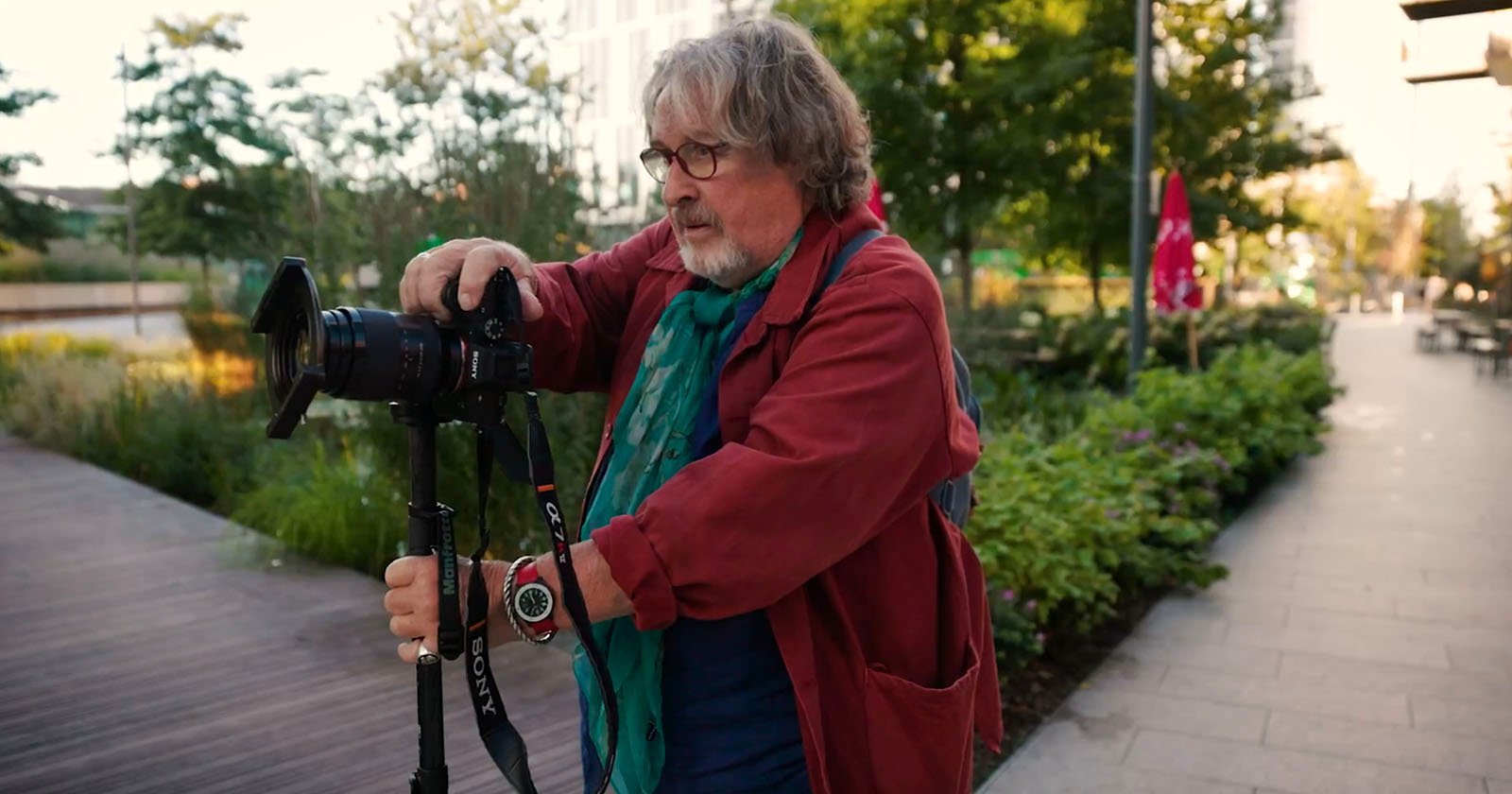 A person in a worn coat and backpack crouches on a city street, aiming a vintage camera. They appear focused on capturing a photo. Other people and blurred buildings are visible in the background.