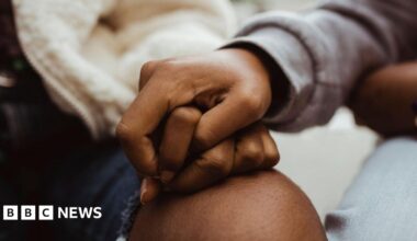 Cropped image of female friends holding hands. The woman on the left is wearing a white zipped bomber jacket with ripped dark denim jeans and the friend on the right has a grey jumper on and light washed denim jeans.