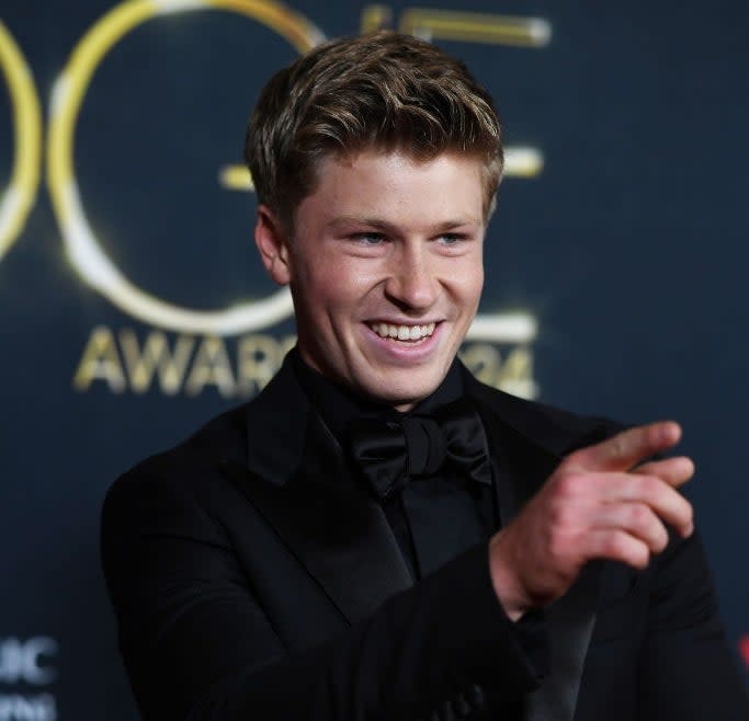 Person in formal black tuxedo smiling and pointing at a media event