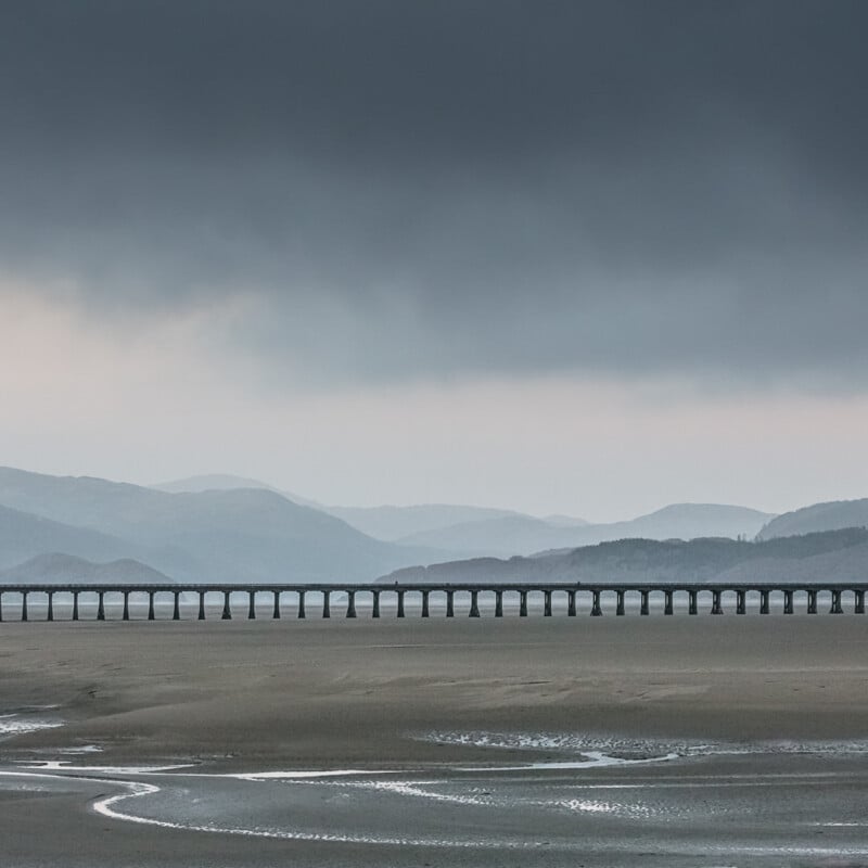 A long railway bridge stretches across a sandy, tidal landscape with distant, misty hills under a moody, overcast sky.