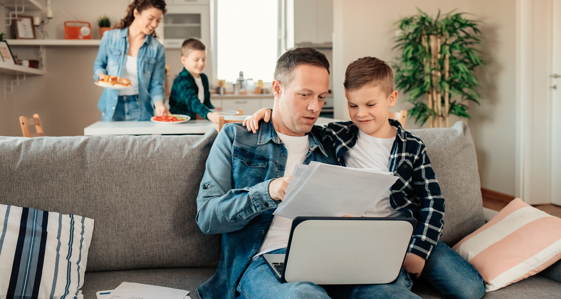 Father Works From Home While His Younger Son Makes Him Company While Mum With Other Son Sets Lunch in the Background.