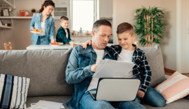 Father Works From Home While His Younger Son Makes Him Company While Mum With Other Son Sets Lunch in the Background.