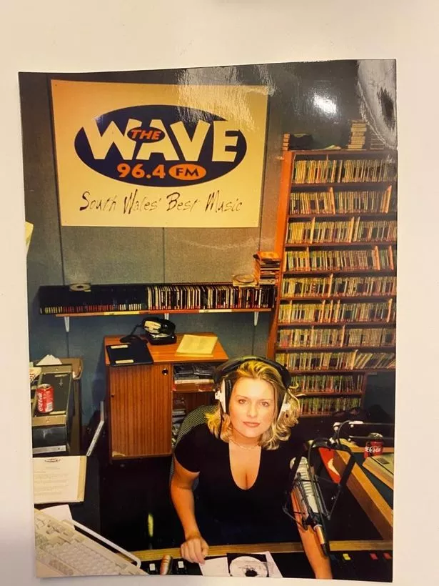 A woman in headphones with CDs behind her and a sign for The Wave radio station