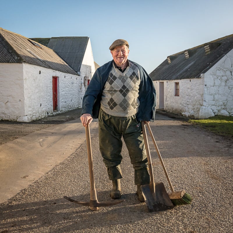 An older man in outdoor work clothes stands smiling between white farm buildings, holding a scythe, shovel, and broom on a sunny day.