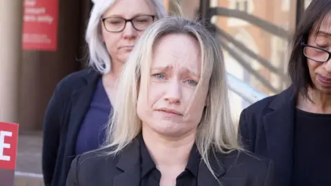 Shaun Whitmore/BBC A woman with blonde hair wearing all black. She looks devastated and is holding back tears. There are people standing behind her also looking very upset.