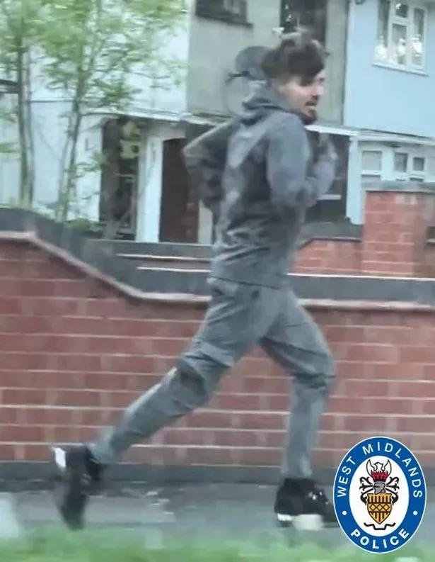 A CCTV image of a man who is running along a pavement and is white, with black hair and a beard, wearing a grey hoodie, grey cargo trousers, black trainers and socks

