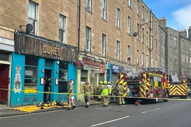 An image of eight firefighters and two fire engines outside business premises, with a hose trained on a doorway with smoke pouring out.
