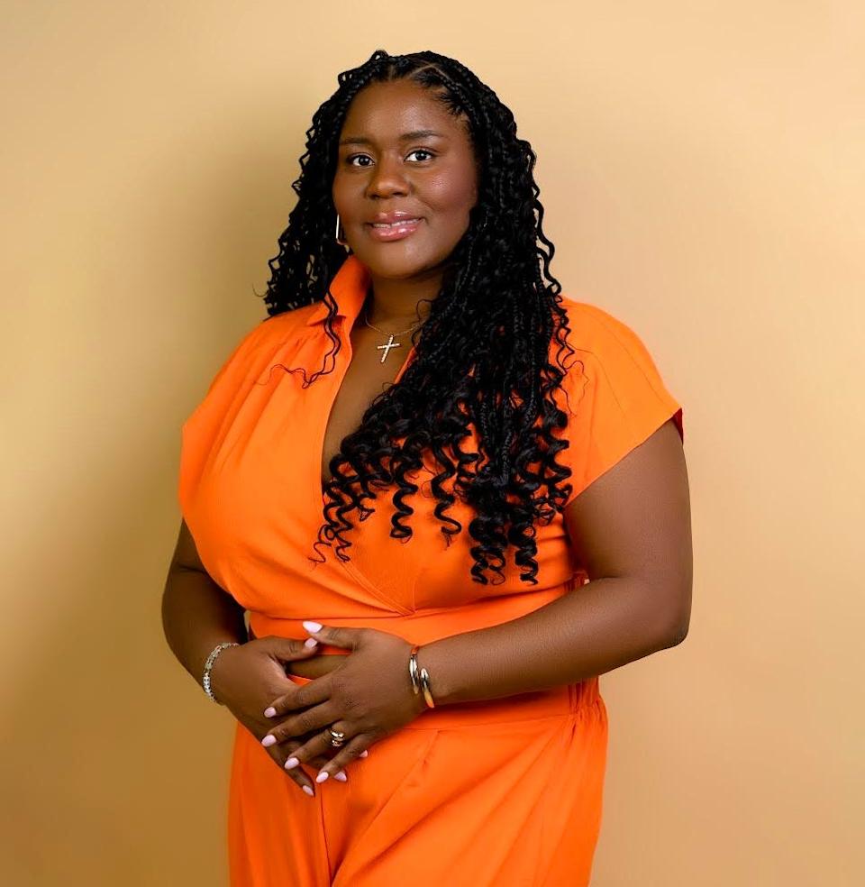 A woman with long, curly braids smiles in a short-sleeved jumpsuit