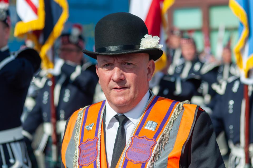 Man in bowler hat and orange sash at a parade.