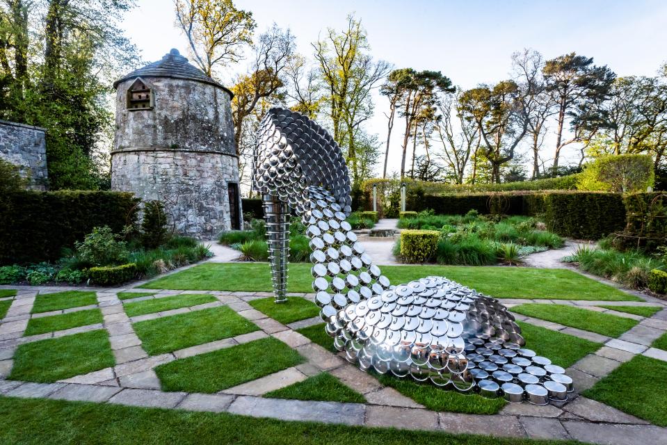 Sculpture of a giant high-heeled shoe made of stacked metallic pots in a garden.