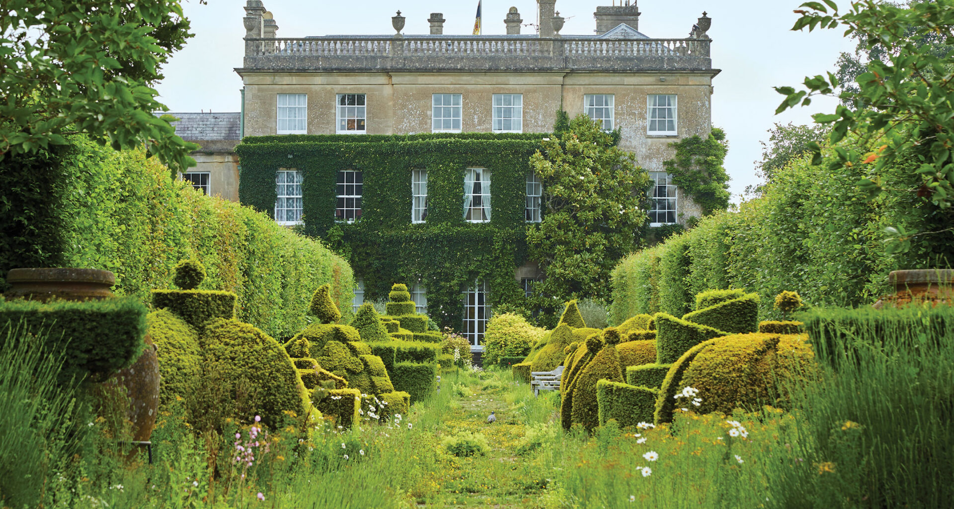 Manor house with lush green gardens, topiary forms, and a variety of flowers, creating a serene and picturesque scene.